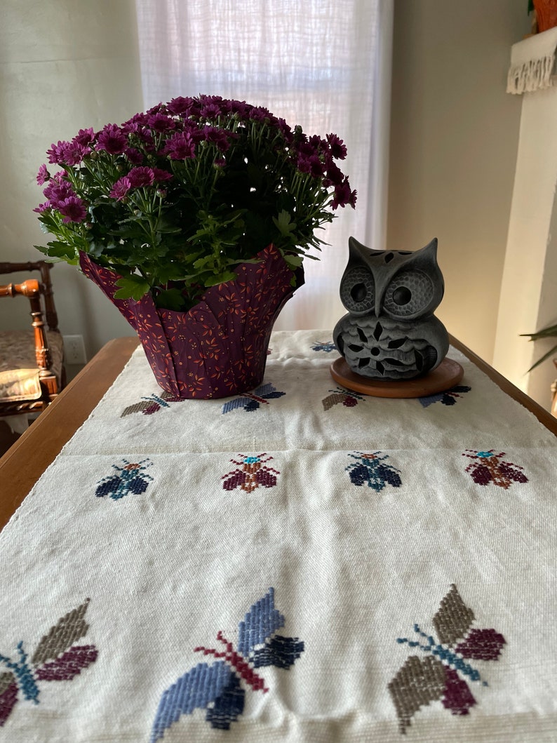 K&ouml;nnte beinhalten: Ein wei&szlig;er Tischl&auml;ufer mit gestickten Schmetterlingsmotiven in Braun-, Lila- und Blaut&ouml;nen. Eine Topfpflanze mit lila Bl&uuml;ten und eine graue Eulenfigur stehen auf dem Tisch.