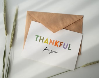 May include: A white card with the word "THANKFUL" in colorful letters and the words "for you" handwritten in black ink. The card is in a brown envelope and there are two stalks of wheat on a white surface.