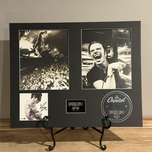May include: A black framed display with three monochrome photographs and a CD. The photographs show a performer on stage, a laughing close-up, and a signed image. The CD is labelled "Capitol" and "IDOLS".
