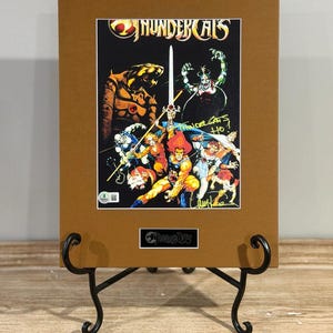 May include: A framed print of the Thundercats cartoon. The artwork features the Thundercats characters with the Thundercats logo at the top. The print is displayed on a black metal stand.