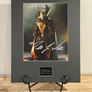 以下が含まれることがあります： ウィル・ジャイ・ホワイトのサイン入り、コミックキャラクター、スポーンの額装プリント。画像は、赤いマントを着た暗いコスチュームのスポーンを、スモーキーな背景に描いています。プリントは黒でマット加工され、装飾的なスタンドに展示されています。