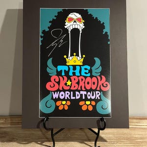 May include: A framed print featuring a cartoon skull with sunglasses and a crown, with the text "THE SK*BROOK WORLD TOUR" in colorful lettering. The print is displayed on a black easel.