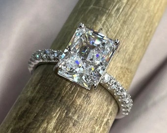 May include: A radiant-cut diamond ring with a silver band. The ring features a large, rectangular-shaped diamond as the centerpiece, with smaller diamonds set along the band. The ring is displayed on a wooden dowel against a soft, pinkish background.
