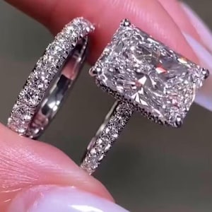 Può includere: Due anelli di diamanti. Uno presenta un grande diamante a taglio rettangolare con incastonatura a alone. Il secondo è una fascia con piccoli diamanti incastonati a pavé. Entrambi gli anelli sono argentati e scintillanti.