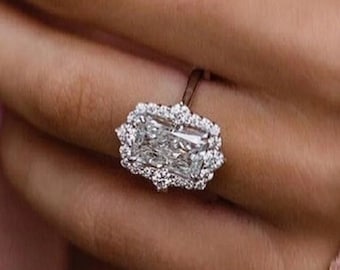 May include: A close-up of a silver engagement ring with a large, rectangular, clear diamond surrounded by smaller diamonds. The ring is on a person's finger.