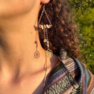 May include: Gold earrings with a triangle shape and a dangling flower charm. The earrings are adorned with pink and white stones.