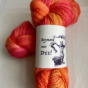 May include: A skein of hand-dyed yarn in shades of orange, yellow, and pink. The yarn is wrapped with a label that reads "Reynard must Dye!" and features a whimsical illustration. Ideal for knitting and crochet projects.
