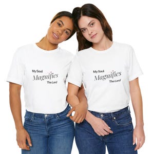 Puede incluir: Dos personas con camisetas blancas y vaqueros azules. Las camisetas tienen el texto "My Soul Magnifies The Lord" en escritura negra. La imagen está bien iluminada y los sujetos sonríen.