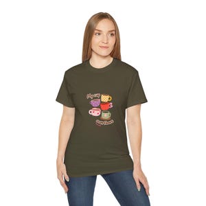 May include: An olive green t-shirt with a design of stacked coffee cups in various colours and patterns, including pink, yellow, and green. The text "My cup overflows" is also visible on the shirt.