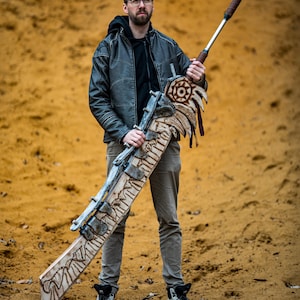 May include: A person holding a large, intricately carved wooden sword with a metal blade and a leather handle. The sword has a decorative design with a skull and crossbones motif. The person is wearing a black leather jacket and jeans.