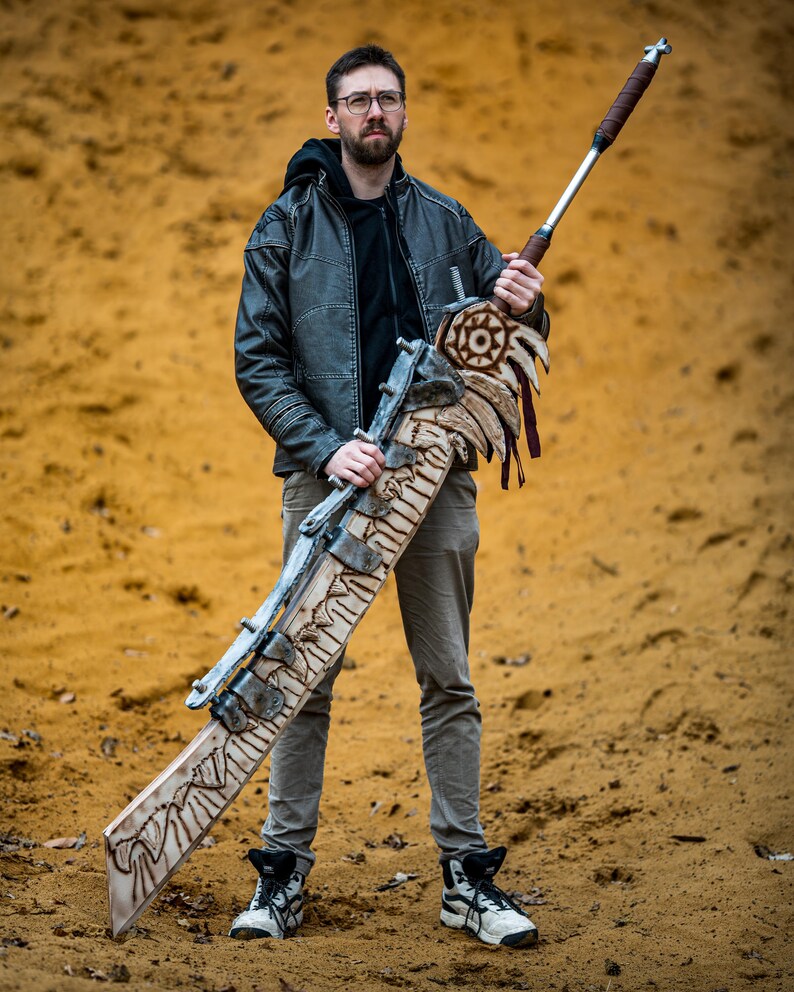 May include: A person holding a large, intricately carved wooden sword with a metal blade and a leather handle. The sword has a decorative design with a skull and crossbones motif. The person is wearing a black leather jacket and jeans.