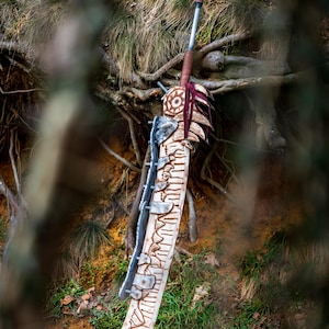 May include: A large, intricately carved wooden sword with a metal blade and a red and brown leather handle. The sword is lying on the ground in a forest setting.