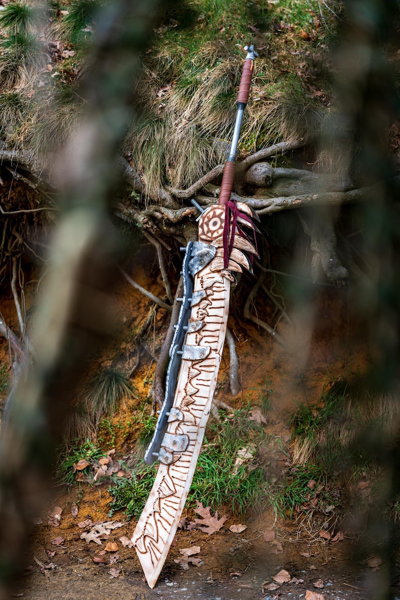 May include: A large, intricately carved wooden sword with a metal blade and a red and brown leather handle. The sword is lying on the ground in a forest setting.