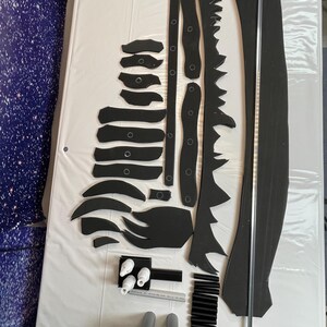 May include: A set of black foam pieces for crafting a dragon costume. The set includes a long, thin piece of foam, a long, curved piece of foam, and several smaller pieces of foam. There are also white plastic pieces, black plastic pieces, and gray PVC pipes.