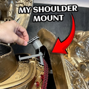 May include: A close-up of a gold-colored shoulder mount with the text "MY SHOULDER MOUNT" in white. A hand is holding a gray piece of the mount. A red arrow points to the mount.