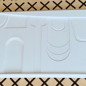 May include: A rectangular white silicone mould featuring various recessed geometric shapes, including arches, vertical lines, and curved segments, designed for casting or moulding materials. The mould has a smooth surface and defined edges.