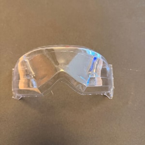 Op de afbeelding: Een helder, plastic beschermingsschild voor de ogen. Het schild heeft een gebogen vorm met een centrale inkeping en is ontworpen om over de ogen te passen. Het materiaal is transparant en lichtgewicht.