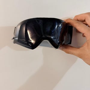 Könnte beinhalten: Schwarze Skibrille mit glänzendem Finish. Die Brille hat eine breite, gewölbte Linse und einen dicken Rahmen. Das Bild zeigt die Brille, die von einer Hand vor einem weißen Hintergrund gehalten wird. Die Brille soll die Augen vor den Elementen schützen.