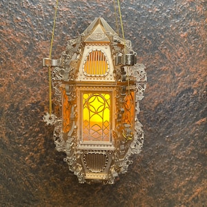 May include: Ornate, gold-colored lantern with intricate detailing, hanging against a textured brown background. The lantern features a geometric design on the glass panels, and emits a warm, yellow glow. The lantern is suspended by a thin, gold-colored cord.