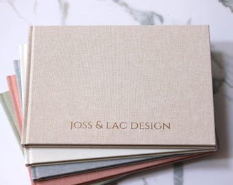 May include: A stack of rectangular photo albums with fabric-textured covers in beige, white, gray, and coral. The top beige album features the text JOSS & LAC DESIGN embossed in a dark brown font on the front cover.