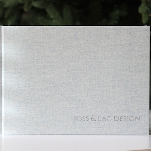 Può includere: Un libro grigio chiaro con una trama di lino e la scritta "JOSS & LAC DESIGN" in rilievo sulla copertina. Il libro è in piedi su una superficie bianca, con un cestino di vimini e del verde sullo sfondo.
