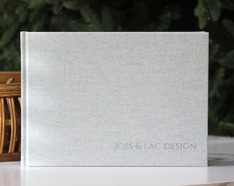 May include: A light gray linen-textured book with the text "JOSS & LAC DESIGN" embossed on the cover. The book is standing upright on a white surface, with a wicker basket and greenery in the background.