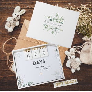 May include: A white gift box with a countdown calendar inside. The calendar has three white squares with the number 0 on each square. The text "DAYS" is below the squares. The text "FROM MISS TO MRS" is below the word "DAYS". The box has a white lid with the text "Let the countdown begin" and a watercolor design of green leaves. The box is on a wooden surface.