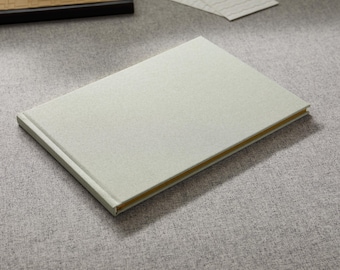 May include: A rectangular hardcover book with a light gray, textured fabric cover. The spine and edges of the pages are visible, showing a gold-colored finish along the side of the book. The book is positioned at an angle on a flat surface.