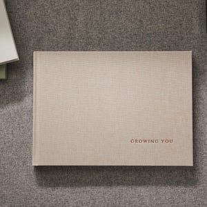 Può includere: Un libro beige rivestito in lino con le parole "GROWING YOU" in lettere rosse. È visibile un altro libro con lo stesso testo, insieme a una penna e un piccolo vassoio. Gli oggetti sono su una superficie grigia strutturata.