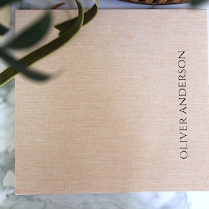 May include: A rectangular, light tan box with the name "OLIVER ANDERSON" printed vertically in black. A green ribbon and leafy branch are in the upper left corner. The box has a textured surface.