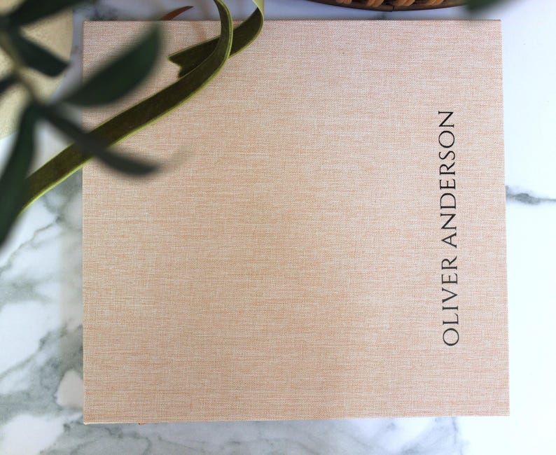 May include: A rectangular, light tan box with the name "OLIVER ANDERSON" printed vertically in black. A green ribbon and leafy branch are in the upper left corner. The box has a textured surface.