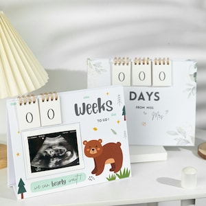 Puede incluir: Un calendario de cuenta atrás blanco con un diseño de oso marrón. El calendario tiene dos pantallas de números abatibles para las semanas y tres para los días. El calendario tiene un marco de fotos con una imagen de ultrasonido y el texto "¡Semanas para ir!" y "¡Apenas podemos esperar!"