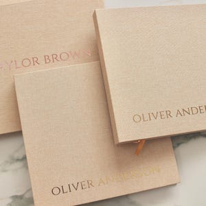 May include: A collection of square, light peach-colored notebooks with embossed text. The text includes the names "TAYLOR BROWN" and "OLIVER ANDERSON" in a metallic rose gold color. The notebooks have a textured fabric cover and a ribbon bookmark.