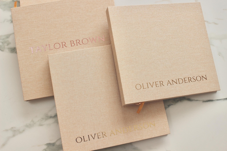 May include: A collection of square, light peach-colored notebooks with embossed text. The text includes the names "TAYLOR BROWN" and "OLIVER ANDERSON" in a metallic rose gold color. The notebooks have a textured fabric cover and a ribbon bookmark.