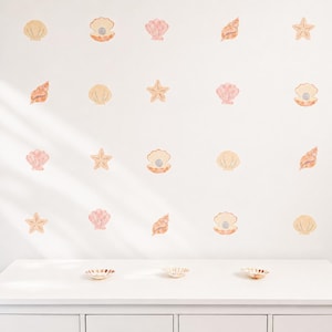 May include: A collection of watercolour-style wall decals depicting seashells and starfish in shades of peach, beige, and tan. The decals are arranged on a white wall, creating a coastal theme. Three small decorative dishes are on a white surface.