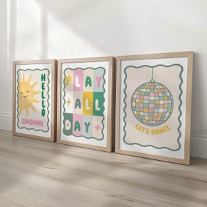 May include: Three framed art prints with a light wood finish. The prints feature retro-style designs: a smiling sun with "Hello Sunshine", a grid with "Play All Day", and a disco ball with "Let's Dance". Each print has a decorative border.