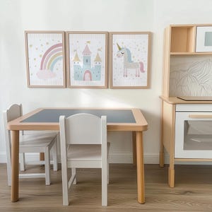 Pu&ograve; includere: Un'area giochi per bambini con un tavolo in legno, piano grigio e due sedie bianche. Sopra, tre stampe incorniciate raffigurano un arcobaleno, un castello e un unicorno. A destra c'&egrave; una cucina giocattolo in legno. Il tavolo &egrave; lungo circa 76 cm.