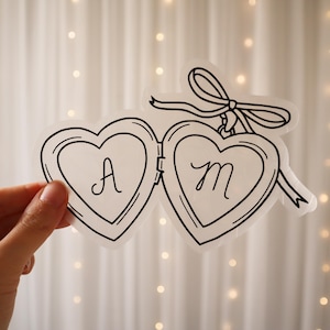 May include: A transparent heart-shaped locket sticker featuring the letters "A" and "M". The locket is outlined in black and has a bow at the top. The sticker is held by a hand against a soft, blurred background.