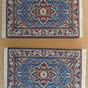 Op de afbeelding: Twee kleine, rechthoekige, witte tapijten met ingewikkelde bloemmotieven in tinten rood, blauw en geel. De tapijten hebben franjes aan de randen en zijn gemaakt van een geweven stof.