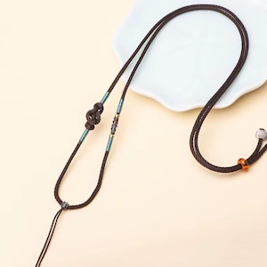 May include: A brown cord necklace with a knotted design and small blue and gold beads. The necklace is on a white surface.