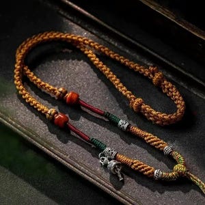 Może przedstawiać: Bransoletka z plecionego sznurka w kolorze brązowym, czerwonym i zielonym z srebrnymi koralikami i srebrnym zawieszką w kształcie dzwonka. Bransoletka ma dwa drewniane koraliki z czerwonymi i białymi akcentami.