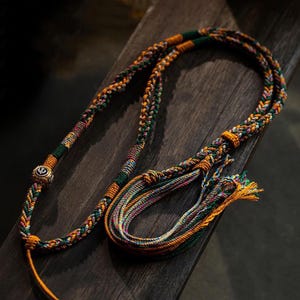 Op de afbeelding: Een veelkleurige gevlochten koordsnoer met een gouden bedel. Het koord is gemaakt van groene, oranje en roze draden. De bedel is een ronde schijf met een ontwerp.