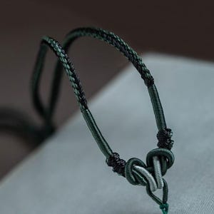 Op de afbeelding: Close-up van een donkergroene en zwarte gevlochten koordketting. De ketting heeft een geknoopt ontwerp met een zilverkleurig metalen accent. Het koord is diepgroen met zwarte accenten, waardoor een getextureerd uiterlijk ontstaat. De achtergrond is wazig.