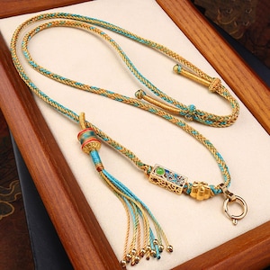 Puede incluir: Un collar de oro y turquesa con un borla y un colgante decorativo. El colgante es de oro y tiene una piedra verde en el centro.