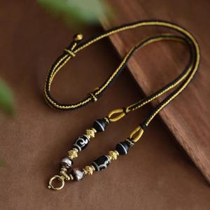 Könnte beinhalten: Eine handgefertigte Halskette mit schwarzem und goldenem Kordel, verziert mit gemusterten schwarz-weißen Perlen und goldenen Akzenten. Die Halskette hat einen goldenen Verschluss und einen verstellbaren Schiebeknoten. Das Design ist aufwendig und elegant.