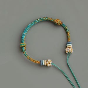 Puede incluir: Un collar artesanal con un cordón trenzado en tonos turquesa, dorado y marrón. El collar tiene nudos decorativos y pequeñas cuentas de colores en los extremos, con cordones turquesa que se extienden desde la parte inferior.