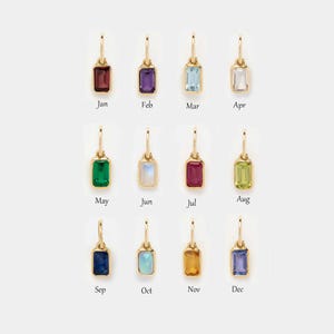 Op de afbeelding: Een verzameling van twaalf goudkleurige oorbellen, elk met een rechthoekige edelsteen in een andere kleur. Elke oorbel is gelabeld met een maand van het jaar, van januari tot december. De edelstenen omvatten rood, paars, blauw en groen.