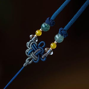 May include: A blue and silver knotted pendant necklace with green and yellow beads. The necklace is made of cord and features a decorative knot design.
