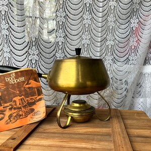 May include: A vintage brass fondue pot with a black knob on the lid, sitting on a wooden table with a booklet titled "et bon appétit" in the foreground.