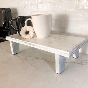 May include: A small, white, distressed wooden riser with four legs. A black mug, a white mug, and a white flower are on top of the riser. The riser is on a white countertop.